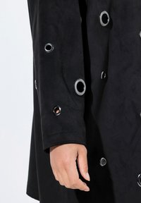 Abrigo de ante negro con acentos de ojales plateados a lo largo de las mangas. Presenta un diseño estructurado y una textura suave. Mano descansando sobre la tela.