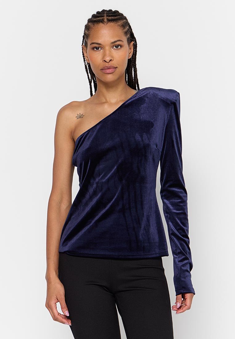 Frau mit geflochtenem Haar, die ein marineblaues Samt-Oberteil mit einem Schulterträger und einem langen Ärmel am rechten Arm sowie schwarze Hosen trägt.