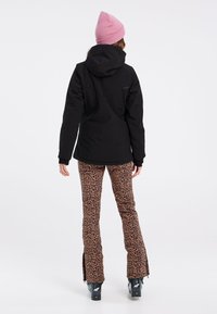Zwarte geïsoleerde jas met een capuchon, gecombineerd met leopardprint flarebroeken en zwarte skibotsen. Roze gebreide beanie op het hoofd.