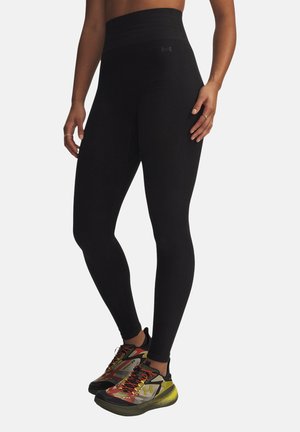 Vrouw draagt zwarte legging met hoge taille en kleurrijke sportschoenen, staand met ontspannen handen langs de zij op een witte achtergrond.