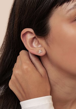 Silberne Ohrstecker in Form kleiner Vögel, verziert mit winzigen Edelsteinen, getragen am Ohr einer Person, mit neutralem Hintergrund und Händen, die das Ohr berühren.