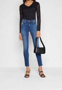 Černá krajková halenka s dlouhým rukávem a výstřihem do V, spárovaná s přiléhavými modrými džíny a černými sandály na podpatku, držící malou černou kabelku.