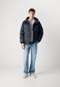 Μπλέ πούφο μπουφάν με κουκούλα, αντίθετους ώμους από καστόρι, ελαστικούς μανικιούς και δύο μπροστινές τσέπες, φορεμένο πάνω από ένα ανοιχτόχρωμο πόλο και τζιν.