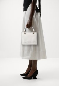 White leather handbag with embossed "VALENTINO" logo, gold hardware accents, and short white handles, held by a person in a white skirt.