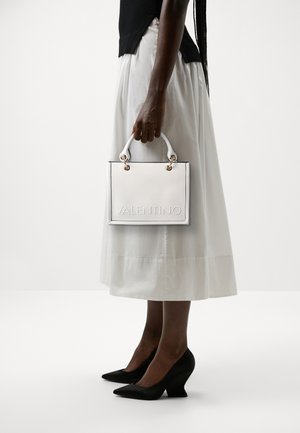 Hvid lædertaske med prægede "VALENTINO"-logo, guldfarvede hardware-detaljer og korte hvide håndtag, båret af en person i en hvid nederdel.