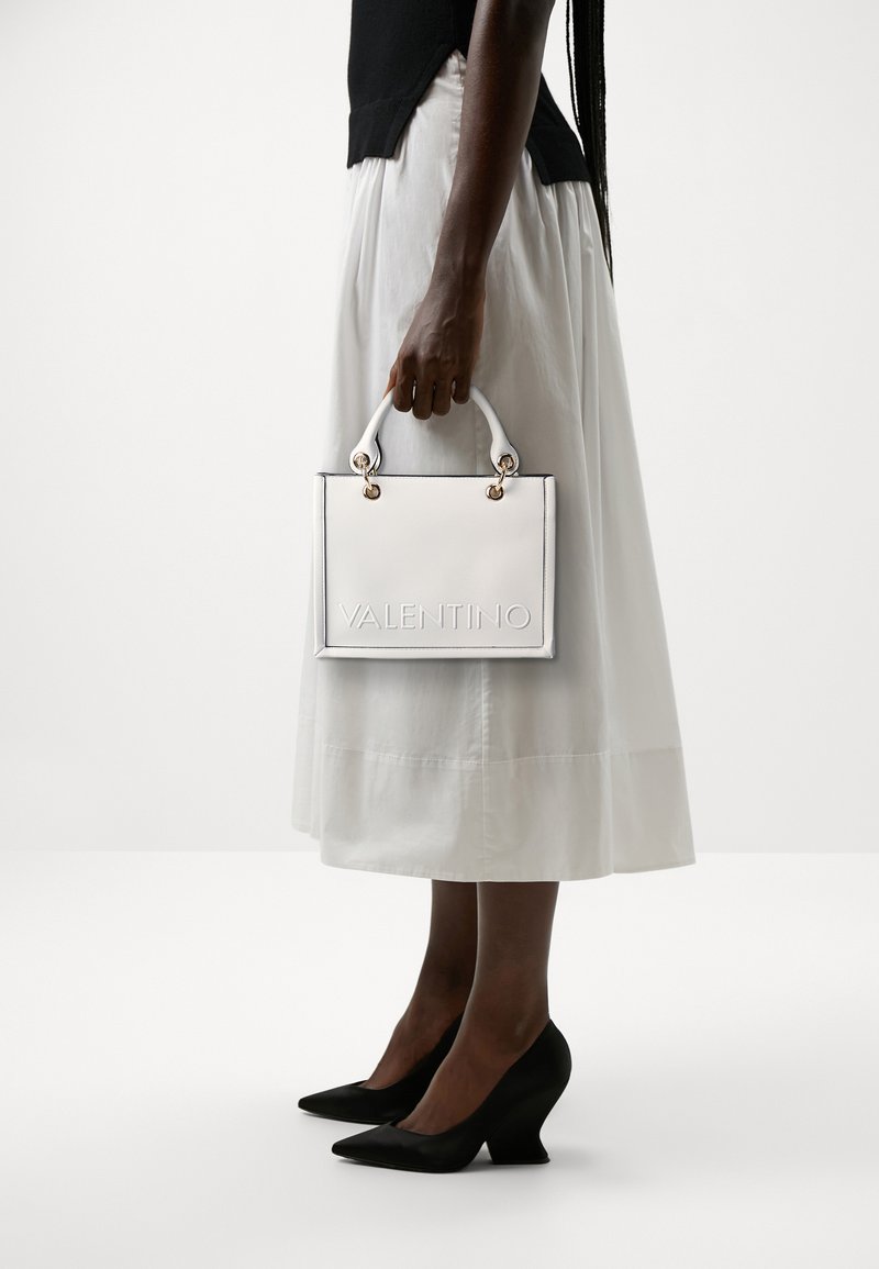 White leather handbag with embossed "VALENTINO" logo, gold hardware accents, and short white handles, held by a person in a white skirt.