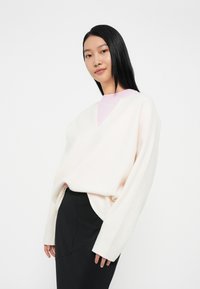 Φωτεινό ροζ oversized πουλόβερ σε μαλακό πλέξιμο με σχέδιο V. Φοριέται πάνω από ένα αντίθετο ροζ μπλουζάκι με στρογγυλή λαιμόκοψη και συνδυάζεται με μια μαύρη φούστα.