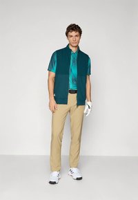 Άνδρας που φοράει πετρόλ πόλο, σκούρο πράσινο γιλέκο χωρίς μανίκια, μπεζ παντελόνι, λευκό γάντι γκολφ και λευκά αθλητικά παπούτσια, στέκεται σε γκρι δάπεδο.