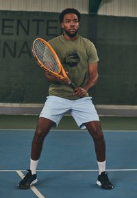 Oranžna teniška lopar, ki ga drži oseba v olivno zeleni majici z grafiko, svetlobluših hlačah in črnih supergah na igrišču.