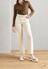 Camisa marrón de manga larga con botones, pantalones crema de talle alto con cinturón y zapatos de plataforma beige gruesos, sobre un fondo claro.