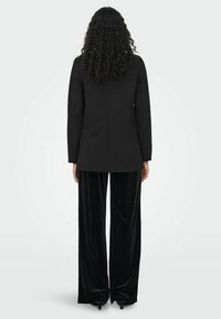 Blazer noir ajusté à manches longues, associé à un pantalon large en velours noir. Texture lisse et design épuré, avec un minimum de détails.