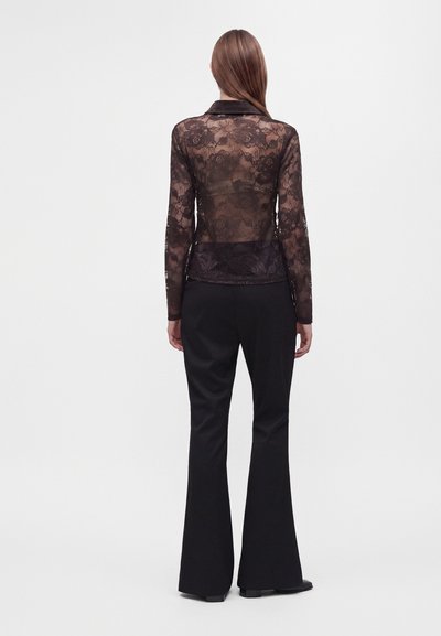 Femme debout, dos à la caméra, portant un haut en dentelle florale noire transparente à manches longues et un pantalon noir évasé, sur un fond uni.