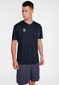 Camiseta deportiva negra de manga corta, con escote en V y logo. Combinada con pantalones cortos gris oscuro que presentan rayas negras a los lados.