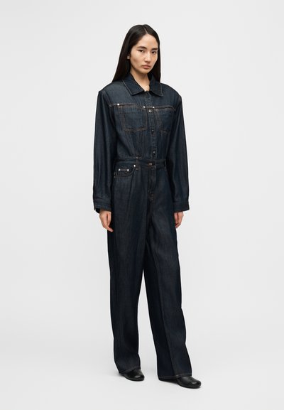 Combinaison en denim foncé avec manches longues, devant boutonné, deux poches poitrine et jambes larges et décontractées. Dotée de coutures contrastantes et d'un col.