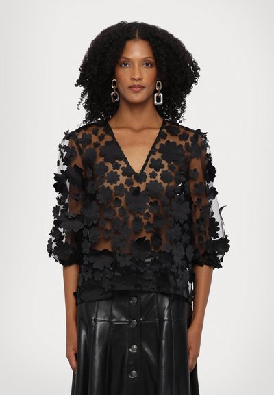 Black sheer blouse featuring 3D floral appliqués, V-neckline, and three-quarter sleeves. Paired with a black leather skirt with buttons.