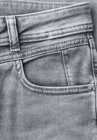 Gros plan sur un jean en denim gris clair montrant la poche avant supérieure, la boucle de ceinture et les détails de couture.