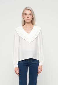 Hvid langærmet bluse med struktureret stof, dyb V-hals og en stor flæsekrave. Parret med blå denimjeans.