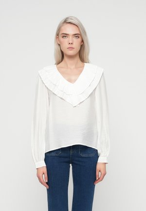 Blouse blanche à manches longues avec un tissu texturé, décolleté en V profond et grand col volanté. Associée à un jean en denim bleu.