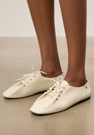 Dunkelhäutige Person trägt cremefarbene, weiche Leder-Schnürschuhe mit quadratischen Zehen und steht auf einem glatten beigen Boden.