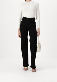 Jeans neri a vita alta abbinati a un top a maniche lunghe bianco a costine. Il top presenta un motivo geometrico e si tiene una piccola borsa beige.