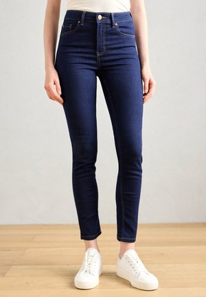 Schmal geschnittene dunkelblaue Jeans, kombiniert mit weißen Sneakers auf Holzfußboden, neutralem Hintergrund und teilweise sichtbarem Oberkörper mit seitlich hängenden Händen.