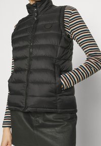Gilet puffer nero con colletto alto, design a righe orizzontali, chiusura con zip e tasche laterali, indossato sopra una camicia a maniche lunghe a righe.