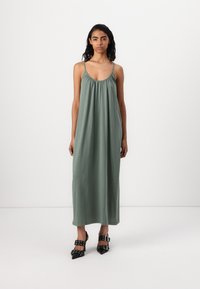 Long, green linen dress with a gathered neckline and thin straps, paired with black pointed-toe heels featuring decorative buckles.