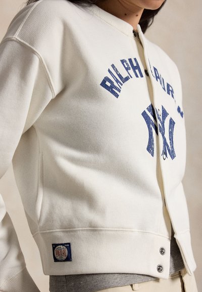 Γυναίκα που φοράει ένα κρεμ φούτερ με φερμουάρ, με μπλε γράμματα «Ralph Lauren» και «NY» και κουμπιά κοντά στο τελείωμα.