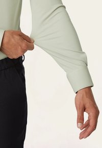 Lange mouwen, lichtgroen shirt met een gladde textuur. Een hand trekt aan de mouw, waardoor een op maat gemaakt manchet detail zichtbaar wordt. Zwarte broek is gedeeltelijk zichtbaar.