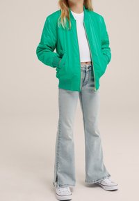 Grön bomberjacka med ribbade ärmar, i kombination med ljusblå utsvängda jeans med sprund i fållen, och vita sneakers.