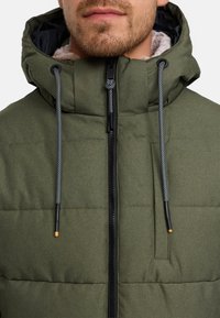 Giacca imbottita verde con colletto alto, cordini regolabili e chiusura zip. L'interno è di colore beige e ha una texture.