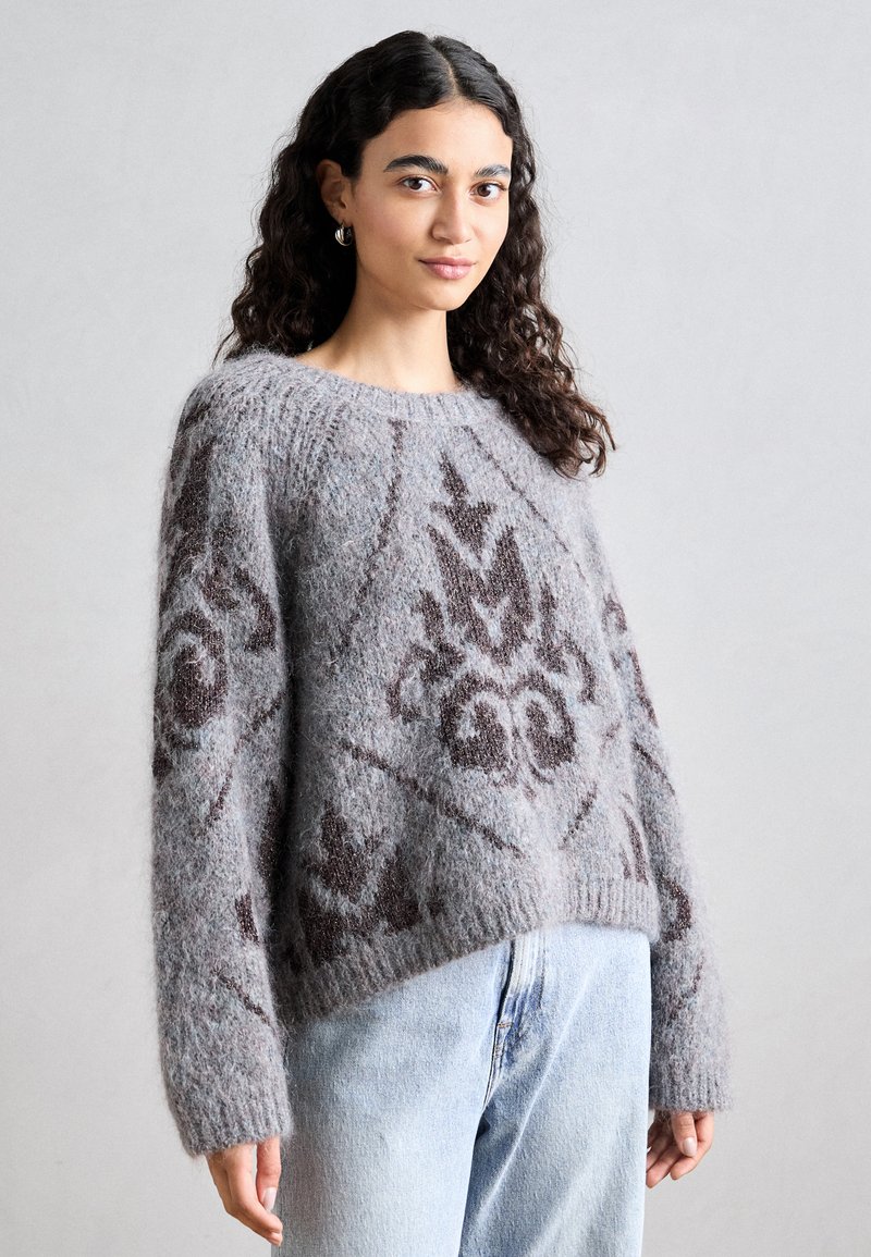Grauer flauschiger Pullover mit braunen, filigranen Mustern, breiten Bündchen und rundem Halsauschnitt, kombiniert mit hellblauen Jeans. Weicher, strukturierter Stoff.