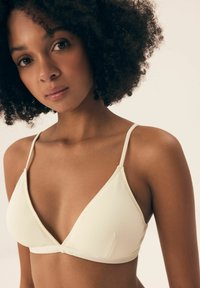 Pale beige triangular bra with thin straps, ribbed texture, and a seamless design, resting on smooth skin. Simple and minimalist style.