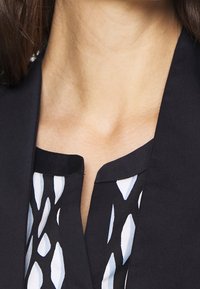 Zwarte blazer met een ronde kraag over een gestreept shirt met blauwe en witte abstracte vormen. Stof lijkt soepel en op maat gemaakt.