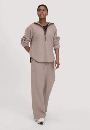Vrouw in een beige fleece pullover met halve rits en bijpassende wijde broek, staande tegen een effen lichte achtergrond.