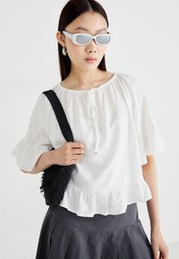Blouse blanche à manches courtes ondulées avec un col à cordon, associée à une jupe plissée gris foncé et un sac à épaule noir texturé.