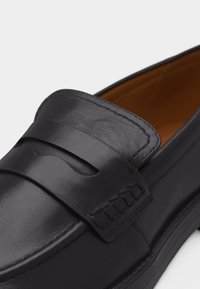 Μαύρο δερμάτινο παπούτσι τύπου penny loafer με λεπτομέρεια κεντημένης λωρίδας και καφέ εσωτερική επένδυση, απεικονιζόμενο σε κοντινό πλάνο.