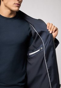 Marineblå jakke med en glat, blank overflade; har hvid pynt og en lynlåslomme indeni. Bæres over en tætsiddende marineblå t-shirt.