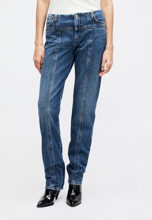 Femme portant un jean taille haute bleu avec des coutures verticales et des bottines à talons aiguilles noires à bouts pointus, debout devant un fond uni.