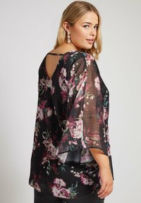 Top floreale nero in tessuto trasparente, con schiena a V, maniche svasate e motivi di fiori colorati in tonalità di rosa e verde.