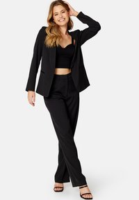 Zwart op maat gemaakt blazer over een zwart cropped top, gecombineerd met op maat gemaakte zwarte broek. Heeft rechte pijpen en een minimalistisch ontwerp.