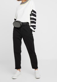 Weißer Strickpullover mit Ärmeln in Navy und Gelb gestreift, schwarze Hose und schwarze Gürteltasche. Weiße Sneakers runden das Outfit ab.