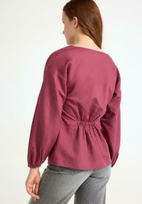 Langærmet skjorte med samlet ryg i dyb mauve stof, der har en bred halsudskæring og voluminøse ærmer, parret med grå bukser.