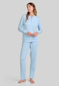 Mujer de pie descalza, vistiendo pijamas de manga larga azul claro con pequeños lunares oscuros, con las manos relajadamente entrelazadas al frente, mirando hacia la derecha.