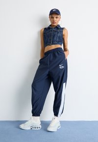 Denim cropped vest, marineblauwe sportieve joggers met witte zijpanelen en logo, en witte sportschoenen met transparante zoolaccenten.