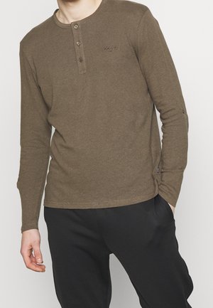 Homme portant une chemise henley manches longues vert olive avec trois boutons et un pantalon noir, une main dans la poche, debout devant un fond blanc.