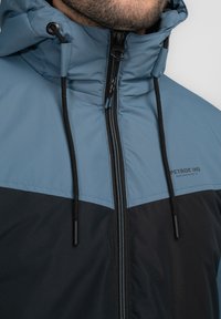 Chaqueta acolchada en azul y negro, con un cuello alto, cordones ajustables y una cremallera frontal con una marca en el pecho.