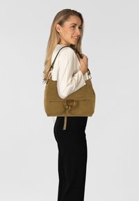 Zandkleurige suède handtas met een nonchalant ontwerp, voorzien van een zwarte leren band en een metalen ringaccent, getoond tegen een neutrale achtergrond.