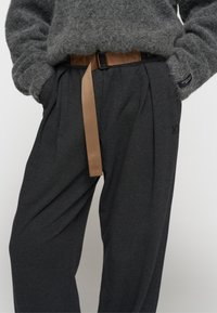 Donkergrijze broeken met een gestructureerde afwerking, voorzien van een bruine riem en voorplooien. Inclusief een label nabij de tailleband.