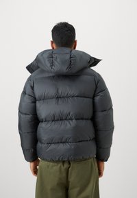 Tamno siva puffer jakna s kapuljačom, s prešanim dizajnom i debelim podstavom za izolaciju. Iza se vidi zaobljena kapuljača i elastični manžeti.